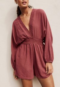 Raspberry-colored romper features a deep V-neck, smocked waist, and long balloon sleeves with gathered cuffs, made from lightweight fabric.