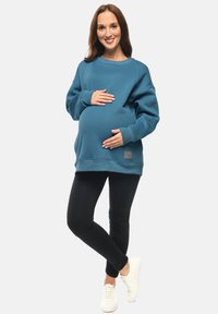 Felpa maternità color teal con scollo rotondo, polsini a costine e logo patch laterale, abbinata a leggings neri e sneakers bianche.