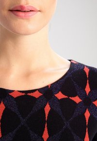 Närbild av en kvinnas nedre ansikte och nacke som bär en svart tröja med ett röd- och mörkbl ått geometriskt mönster.