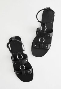 Černé kožené sandály s designem s více popruhy, kulatými kovovými detaily a popruhem přes kotník. Plataná podrážka s matným povrchem.