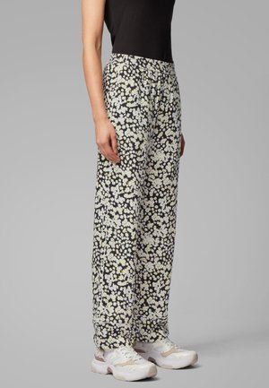 Femme portant un haut noir et un pantalon noir ample avec un imprimé floral marguerite blanche et des baskets blanches, debout devant un fond gris uni