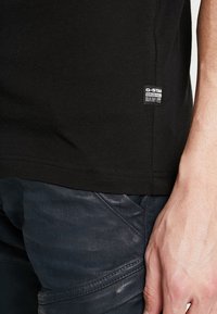 Camiseta negra de algodón con mangas cortas, con dobladillo cosido y una pequeña etiqueta de G-Star cerca del borde inferior en el lado izquierdo.