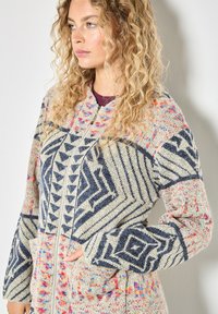 Sweter zapinany na zamek w kolorze beżowym z niebieskimi geometrycznymi wzorami i wielokolorowymi akcentami. Ma kieszenie oraz wysoki kołnierz.