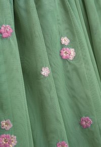 Grüner Tüllstoff mit floralem Stickmuster. Enthält rosa und weiße Blumen mit goldenen Akzenten, die ein strukturiertes, geschichtetes Erscheinungsbild erzeugen.