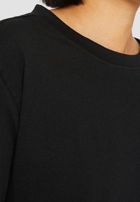 Gros plan sur le cou et l'épaule d'une personne portant un t-shirt noir uni à col rond, avec de courts cheveux foncés visibles.