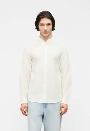 Νέος άνδρας με μεσαίου μήκους σκούρα μαλλιά, ντυμένος με άσπρο πουκάμισο με γιακά μανταρίνι και ανοιχτό μπλε τζιν, στέκεται μπροστά σε απλό λευκό φόντο.