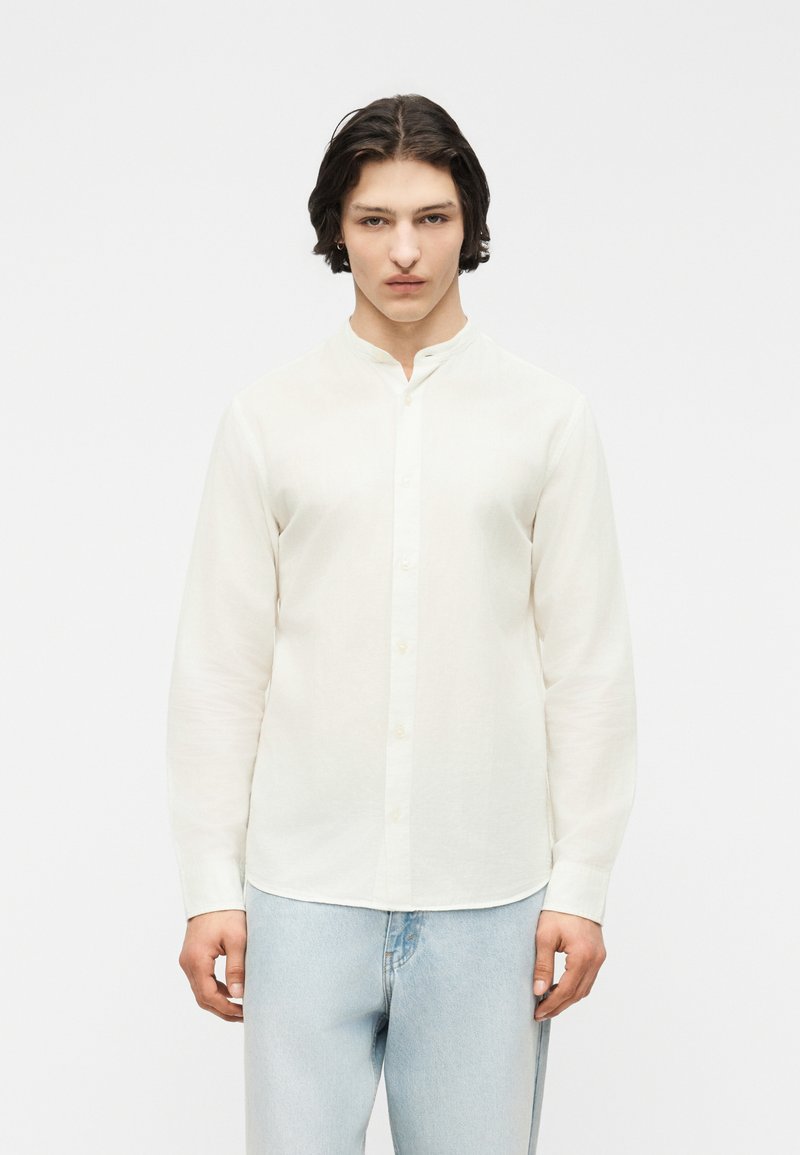 Νέος άνδρας με μεσαίου μήκους σκούρα μαλλιά, ντυμένος με άσπρο πουκάμισο με γιακά μανταρίνι και ανοιχτό μπλε τζιν, στέκεται μπροστά σε απλό λευκό φόντο.