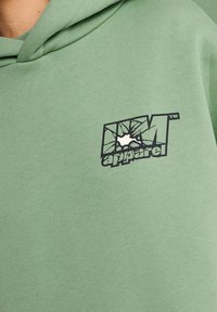 Sudadera verde con capucha, que presenta un logo negro que se asemeja a vidrio roto, impreso en la parte frontal. Textura de algodón suave.
