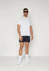 Άνδρας που στέκεται με λευκό αθλητικό μπλουζάκι, μαύρες βερμούδες, λευκές κάλτσες, λευκά αθλητικά παπούτσια και μεγάλες μαύρες αθλητικές ηλιο sunglasses σε ουδέτερο φόντο.