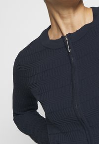 Námořnický modrý zipový kardigan s žebrovanou texturou. Má zaoblený límec a slim fit střih, který ukazuje horizontální a vertikální vzory.