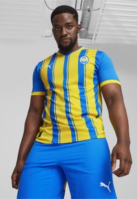 Gul og blå stribede atletiktrøje med korte ærmer og v-hals, med et logo og hvide detaljer, parret med blå atletikshorts.