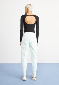 Persoon met kort blond haar, gekleed in een zwart langemouwen top met een grote uitsnede op de rug en lichtblauwe sweatpants, staande op een blauwe vloer.
