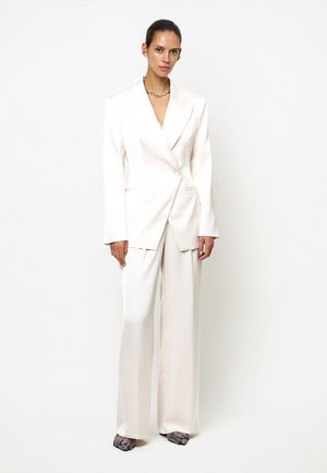 Vrouw draagt een witte oversized blazer en wijde broek met donkergrijze open schoenen tegen een effen witte achtergrond.