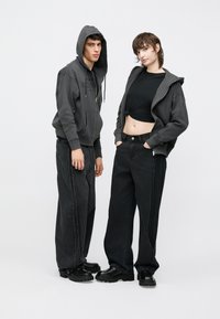 Doi indivizi care poartă ținute coordonate: hanorace cu fermoar gri închis, tricou negru scurt și blugi negri cu picioare largi și cusături contrastante.