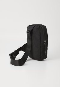 Sac bandoulière noir de forme rectangulaire, avec fermeture éclair et bandoulière réglable. Fabriqué en tissu durable avec une surface texturée.