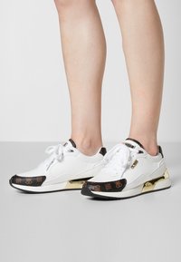 Des sneakers blancs avec des accents noirs à motifs, des finitions dorées et un design bas. Ils présentent des textures contrastées et une pointe ronde.