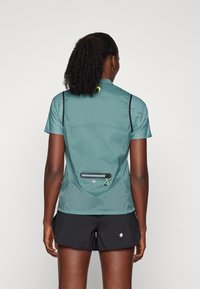 Kurzärmeliges Lauftop in Petrol mit Netzpaneelen, einer Reißverschlusstasche auf der Rückseite und schwarzer Einfassung. Kombiniert mit schwarzen Shorts mit kontrastierendem Akzent.