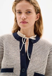 Knitted cardigan in cream and navy with a textured pattern, featuring a ruffled collar, buttons, and pockets.