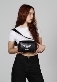 Bolso cruzado sintético negro con cierre de cremallera, logo plateado y diseño de alas, acentos de tachuelas y correa ajustable.