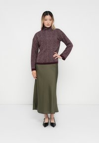 Woman standing with hand on hip, wearing a maroon chevron-patterned sweater, olive green skirt, and black pointed shoes.
