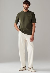 T-shirt di cotone verde oliva con maniche corte e taschino sul petto, abbinata a pantaloni a gamba larga color crema e sneakers beige con dettagli marroni.
