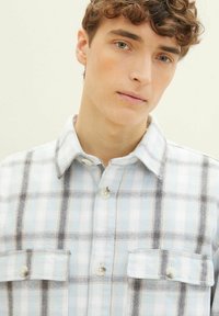 Junger Mann mit lockigem Haar, der ein hellblaues und weißes karriertes Button-up-Hemd trägt und direkt in die Kamera schaut, vor einem schlichten Hintergrund.