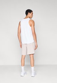 Valkoinen hihaton toppi, beiget shortsit, valkoiset sukat ja vaaleat lenkkarit. Malli seisoo selin yksinkertaisen taustan edessä.