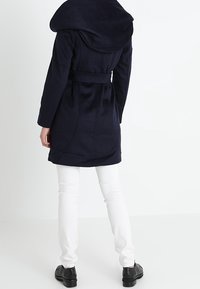 Marinblå kappa med stor luva, omlottbälte och sidofickor. Sammanställd med vita slim-fit byxor och svarta skor. Mjuk textur.