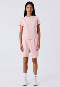 Mujer de pie vestida con una camiseta de manga corta rosa claro y pantalones cortos a juego, con calcetines blancos y zapatillas gruesas blancas.