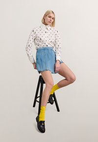 Balta marškinius su gėlėmis, šviesiai mėlynos džinsinės sijonas, geltonos kojinės ir juodi bateliai. Modelis sėdi ant juodo suolo.