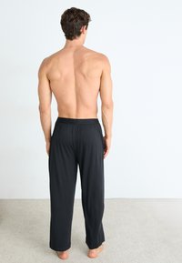 Homme debout pieds nus sur un tapis, tourné vers l'éloignement, portant un pantalon de pyjama noir ample, torse nu contre un fond blanc uni.