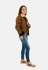 Giacca bomber marrone con colletto in pelliccia sintetica, polsini neri e etichetta arancione; jeans skinny blu; sneakers beige e borgogna.