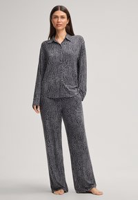 Vrouw die blootsvoets staat, gekleed in een losse zwart-grijze gemusterde blouse met lange mouwen en bijpassende wijde broek voor een effen achtergrond.