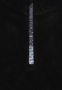 Tela de forro polar negra con una tira reflectante vertical que presenta patrones geométricos de triángulos y texto parcialmente visible cerca de una cremallera.