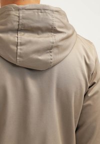 Krémová bundu s kapucí zobrazená ze zadní strany, která zdůrazňuje prošitou kapuci a hladkou texturu látky.