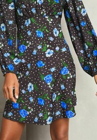 Schwarzes Kleid mit langen Puffärmeln, das mit blauen Blumendrucken und weißen Polka-Dots versehen ist. Leichtes Material mit tailliertem Schnitt und schwingendem Saum.