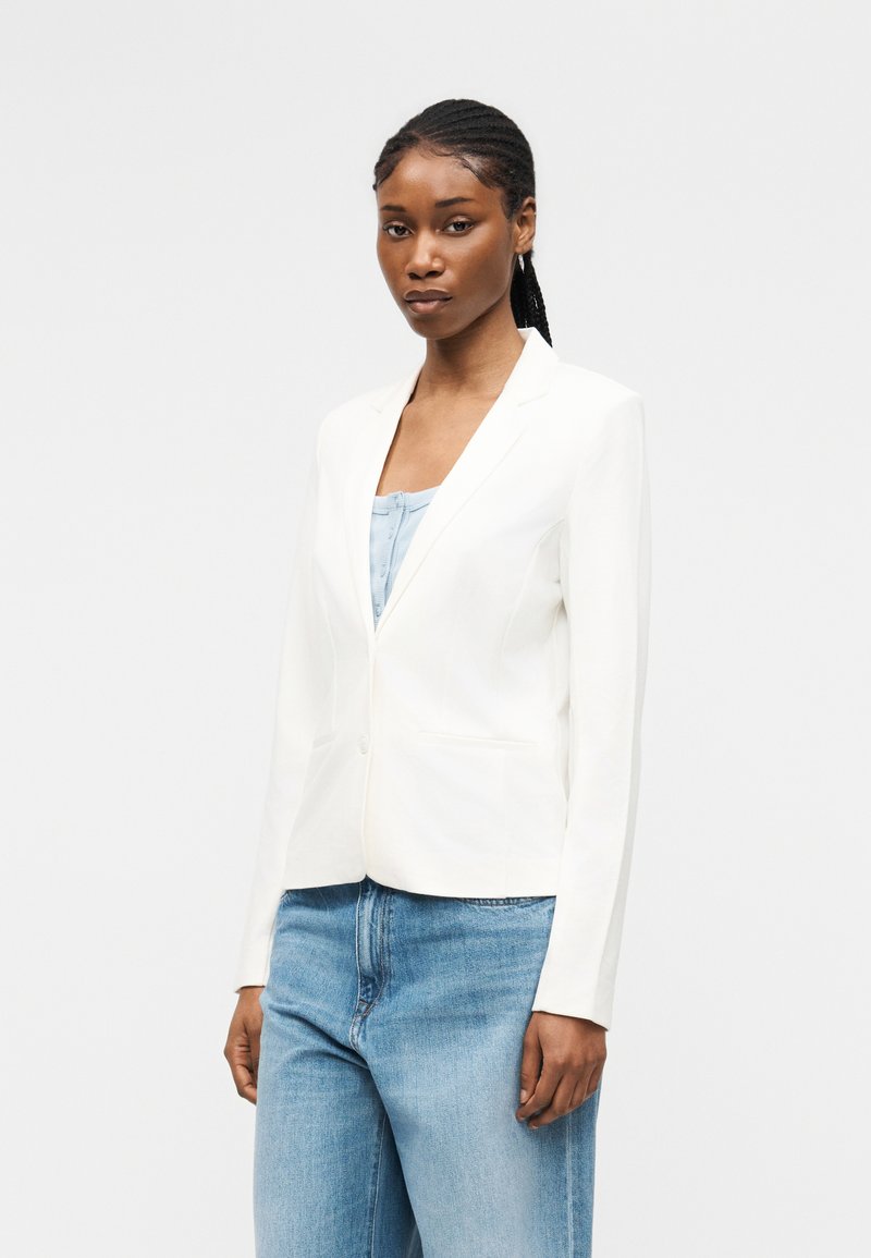 Femme avec une coiffure en tresses cornrows, portant un blazer blanc ajusté et un jean bleu clair, debout devant un fond blanc uni.