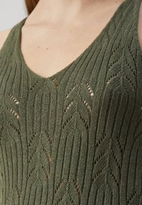 Grønn strikket topp med v-halsdesign, bladmønster og åpenhetsdetaljer, som fremhever teksturert stoff og intrikate sømmer.