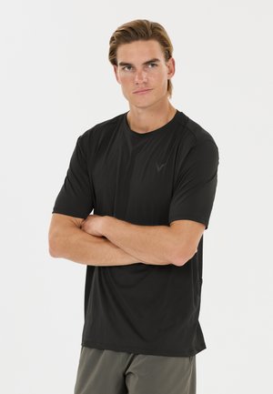 Homme aux cheveux châtain clair portant une chemise noire à manches courtes et un pantalon gris, debout les bras croisés devant un fond blanc.