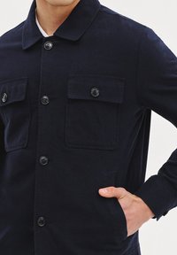 Granatowa kurtka wykonana z tkaniny o teksturowanym wykończeniu. Posiada kołnierz, dwie kieszenie na piersiach z klapkami oraz zapięcie na guziki z przodu.