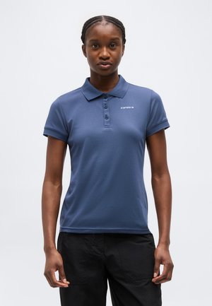 Femeie cu părul împletit, purtând un tricou polo bleumarin cu mânecă scurtă și pantaloni negri, privirea îndreptată înainte pe un fundal simplu.