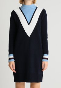 Robe en tricot bleu marine avec un décolleté en V, ornée d'accents blancs et bleu clair. Poignets côtelés en bleu, blanc et bordeaux. Manches longues.