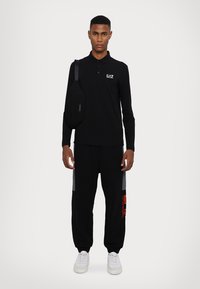 Polo shirt nero a maniche lunghe con logo bianco, abbinato a pantaloni sportivi neri con dettagli grigi e arancioni. Scarpe da ginnastica bianche completano l'outfit.