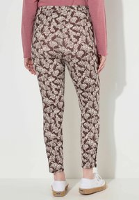 Donna che indossa pantaloni marroni aderenti con motivi a foglia bianchi, abbinati a sneakers espadrilles bianche e una maglietta a maniche lunghe rosa, in piedi contro un muro.