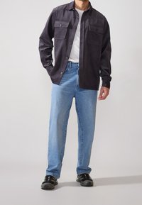 Giacca camicia in velluto a coste nera con due tasche sul petto, indossata sopra una maglietta bianca, abbinata a jeans azzurri chiari e scarpe da ginnastica nere.
