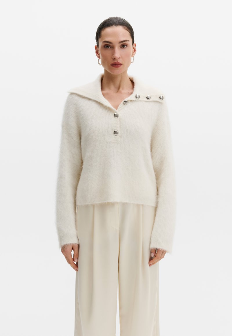 Femme portant un pull blanc duveteux avec des boutons argentés sur le col et le devant, assorti à un pantalon ample de couleur claire.