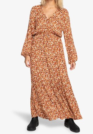Kobieta ubrana w długą sukienkę z długim rękawem, sięgającą do kostek, w kwiatowy wzór w odcieniach pomarańczowego, brązowego i białego, zestawioną z czarnymi botkami do kostek.