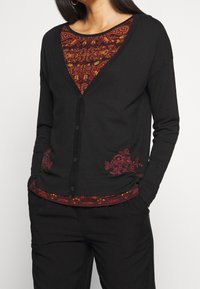 Schwarze Strickjacke mit V-Ausschnitt und roter floraler Stickerei am Saum, über einem rot gemusterten Oberteil getragen. Weicher Stoff, Knopfverschluss.