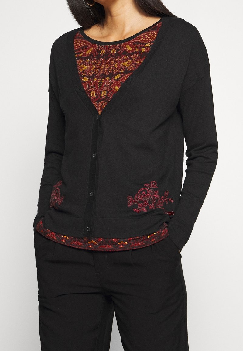 Schwarze Strickjacke mit V-Ausschnitt und roter floraler Stickerei am Saum, über einem rot gemusterten Oberteil getragen. Weicher Stoff, Knopfverschluss.