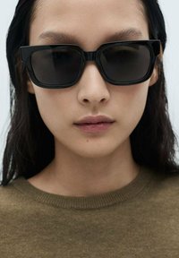 Schwarze rechteckige Sonnenbrille mit dickem Rahmen, dunklen Gläsern und polierter Oberfläche. Das Modell trägt einen olivgrünen Pullover.